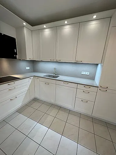 Elegante Häcker-Landhausküche mit Ravenna-Lackfronten in Weiß. Klassisches Rahmen-Design, Porzellan-/Messinggriffe, hochwertige Arbeitsplatte im Stein-Dekor. Inklusive Backofen auf ergonomischer Höhe, Spüleneinheit mit edler Armatur und viel Stauraum.