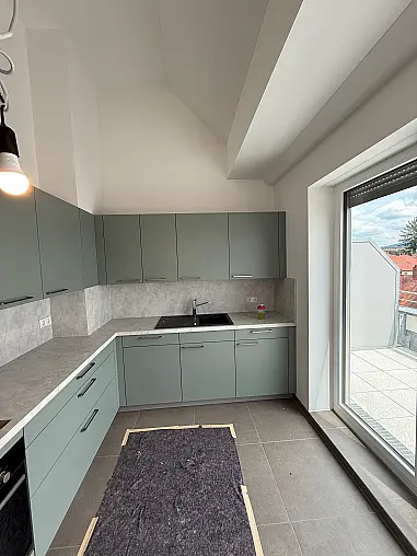 Zeitlos moderne L-Küche mit mattgrünen Fronten und klaren Linien. Die großzügige Arbeitsfläche in Steinoptik harmoniert perfekt mit den eleganten Edelstahlgriffen.