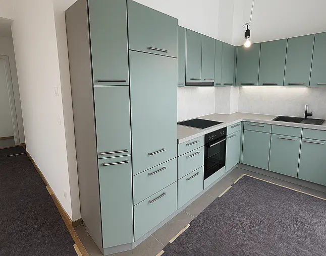 Zeitlos moderne L-Küche mit mattgrünen Fronten und klaren Linien. Die großzügige Arbeitsfläche in Steinoptik harmoniert perfekt mit den eleganten Edelstahlgriffen.