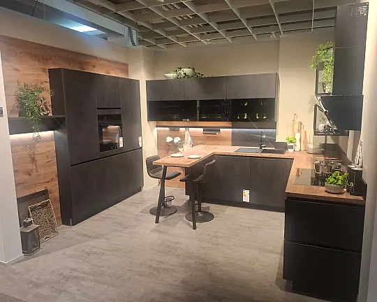 Moderne, elegante L-Küche mit wohnlicher Spachtelbetonoptik in Schwarz, kombiniert mit warmen Alpineiche-Akzenten und hochwertiger Lichtgestaltung. - Typbezeichnung / Modellname: Comet GL - schwarz Spachtelbetonoptik / Alpineiche (Programm T559) -Koje 088-