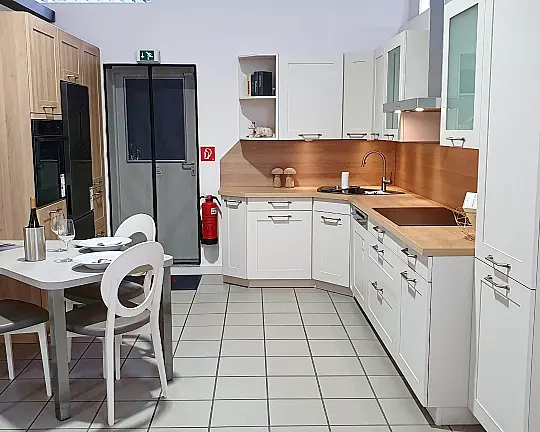 Müsterküche im modernen Landhaus-Stil für gemütliches Ambiente, raffinierte Tischlösung inklusive, L-Form mit Eckspüle und Hochschrankblock mit umbautem Standkühler. - Modell: FRAME, Farben: Magnolia mit Indian Oak,